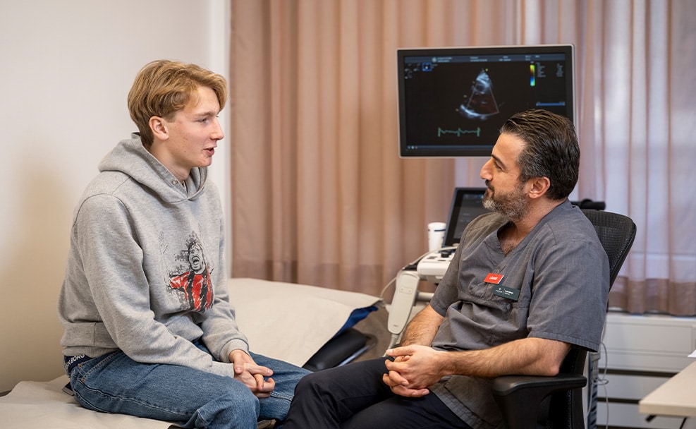 Ted sitter på en undersökningsbänk och pratar med en ansvarig för hjärtscreening på sports cardiology. I bakgrunden ser vi en screening på en dator.