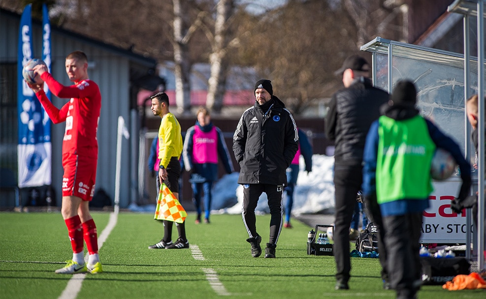 Sidan av en fotbollsplan, en spelare förbereder sig för inkast och tränaren Lukas observerar.